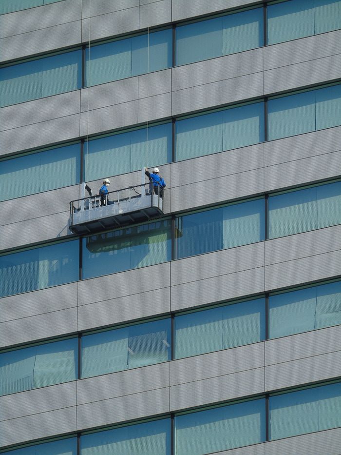 Two employees wash windows on a contemporary glass building exterior while standing on a suspended platform.